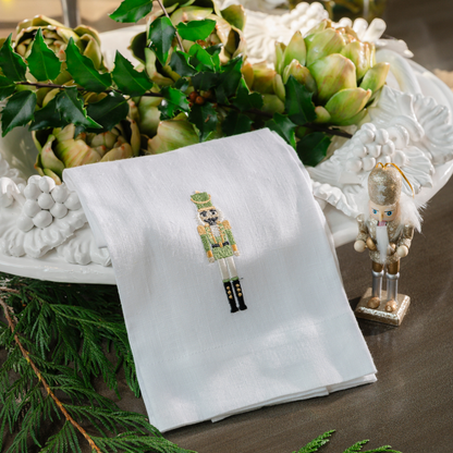 A white tea towel embroidered with a nutcracker in a green uniform laying on a scavo vineyard bowl with artichokes and holly