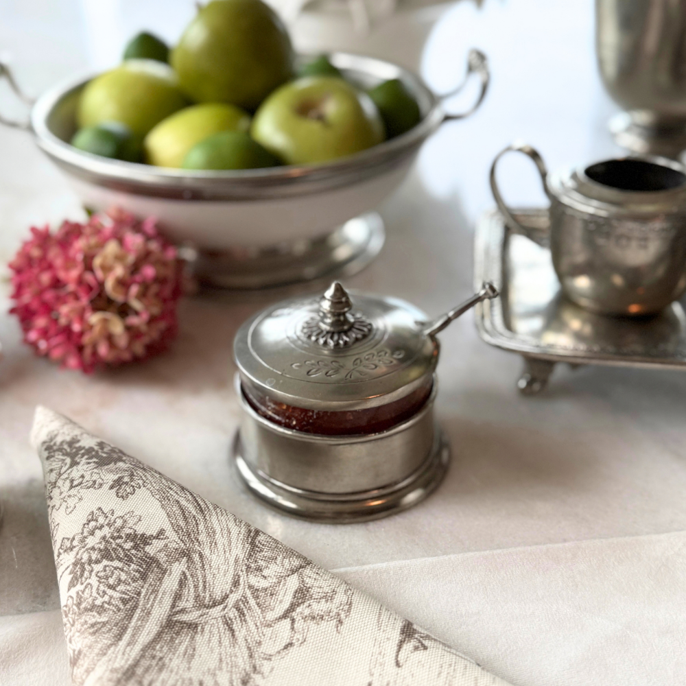 Peltro condiment dish with spoon on a table with fruit in the background and a toile napkin