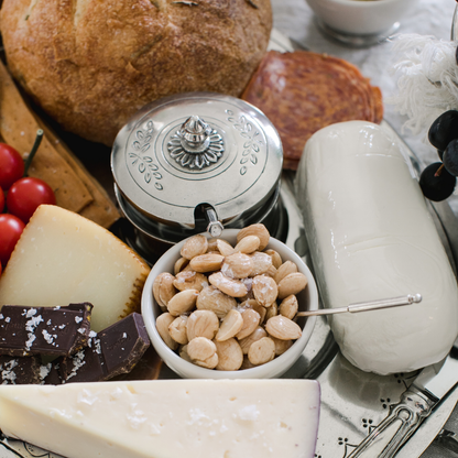 Top view of the Peltro pewter lid condiment dish with spoon on a charcuterie board with nuts, bread, cheese and chocolate