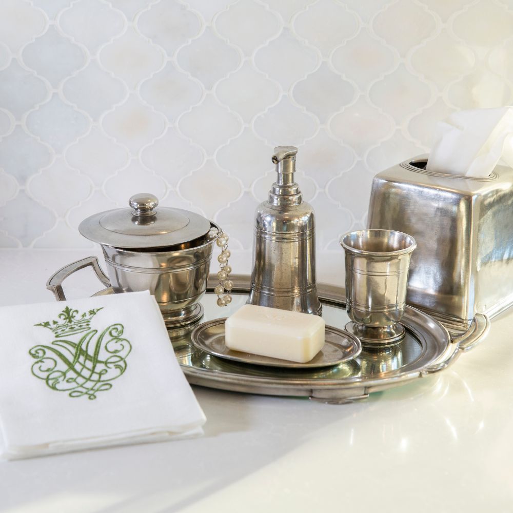 Pewter high end bathroom accessories including a soap dispenser, cup, and tray on a white countertop with a tiled backsplash.