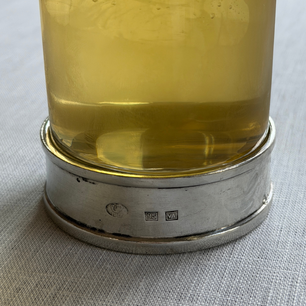 Close-up of the handcrafted Italian pewter base of the Arte Italica Portofino Highball Glass, showing artisan hallmark stamps, filled with a golden cocktail