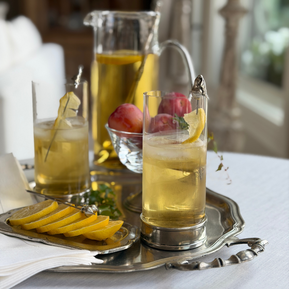 Two Arte Italica Portofino Highball Glasses filled with a golden pear cocktail on ice, styled on a pewter tray with lemon slices, cocktail picks, fresh peaches, and a Portofino glass pitcher, on a white linen tablecloth