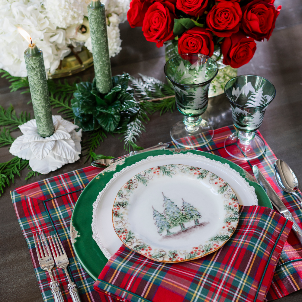 A festive holiday table setting featuring a Natale plate with a Christmas tree illustration on the rim, layered over a green charger plate, a red and green plaid napkin, and matching plaid placemat. The scene includes green candles, red roses in a vase, and green glassware on a dark tabletop.