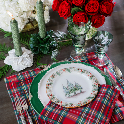 A festive holiday table setting featuring a Natale plate with a Christmas tree illustration on the rim, layered over a green charger plate, a red and green plaid napkin, and matching plaid placemat. The scene includes green candles, red roses in a vase, and green glassware on a dark tabletop.