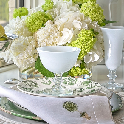 Rialto hand painted Italian Glass Compote in white with Flute and ceramic plates and embroidered linen Napkin on a table with flowers