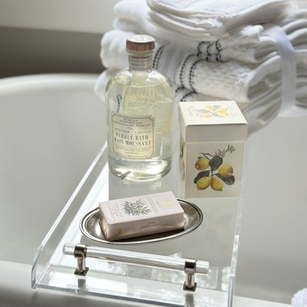 Bottle of soap and a bar of soap on a pewter tray  on a bath caddy with a white towel in the background.