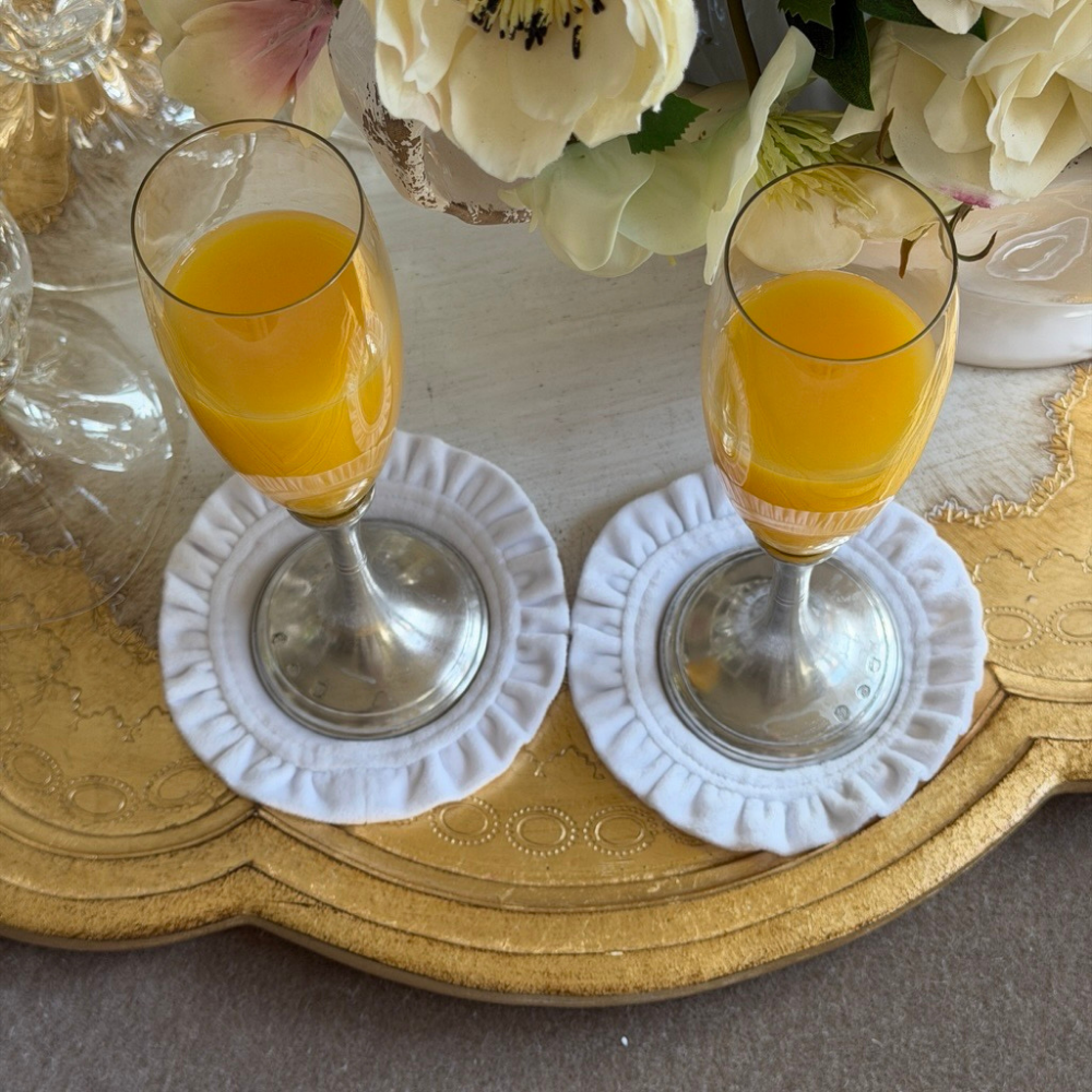 Two Verona flute glasses filled with Orange juice on white round velvet coasters on a gold Florentino tray with flowers