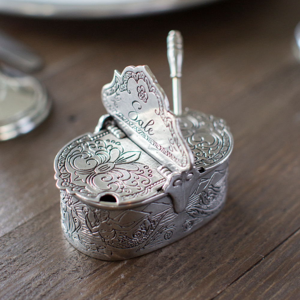 Decorative pewter salt and pepper cellar with intricate designs on a wooden surface