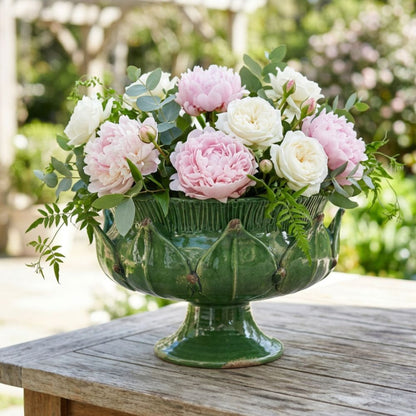 A green ceramic Savo pedestal bowl inspired by an artichoke filled with a floral arrangement of pink and white peonies and green foliage, displayed on a rustic wooden table in an outdoor garden setting.