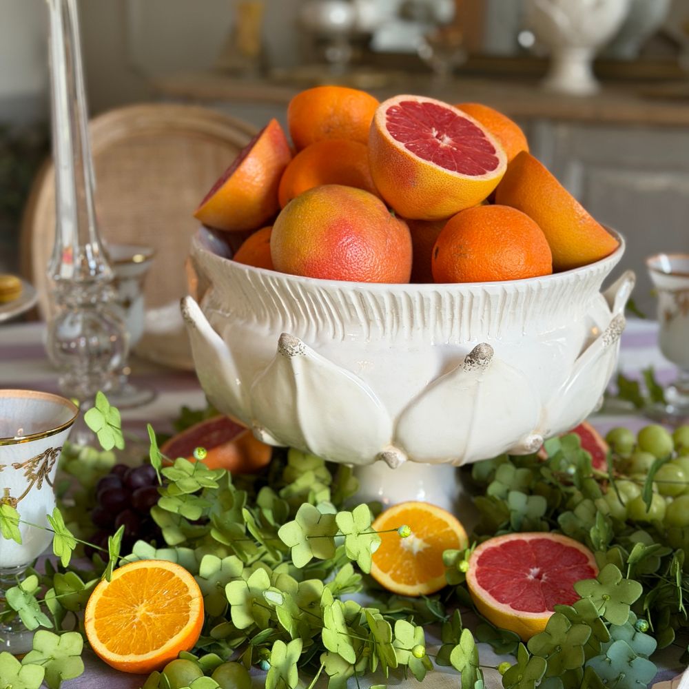 A white ceramic Scavo pedestal bowl inspired by an artichoke filled with citrus fruits, styled on a table with green leaves, sliced oranges, grapes, and glassware in a softly blurred indoor setting.