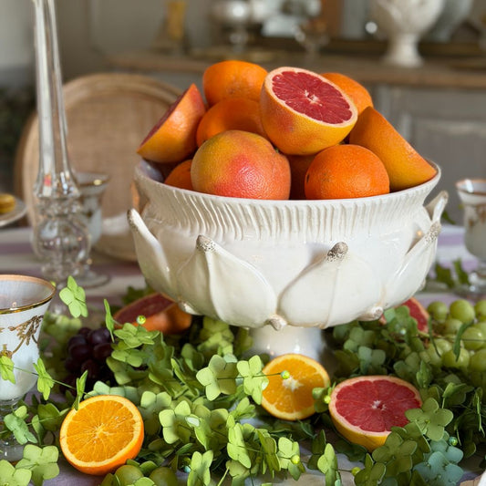 A white ceramic Scavo pedestal bowl inspired by an artichoke filled with citrus fruits, styled on a table with green leaves, sliced oranges, grapes, and glassware in a softly blurred indoor setting.