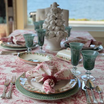 Arte Italica Scavo Fruit Tree Topiary as a centerpiece on a holiday table set with a red Montefalco toile cotton tablecloth, Natale dinner plates, green Scavo wine glasses, Montefalco toile napkins with red beaded napkin rings, and Italian pewter flatware, with a water view in the background
