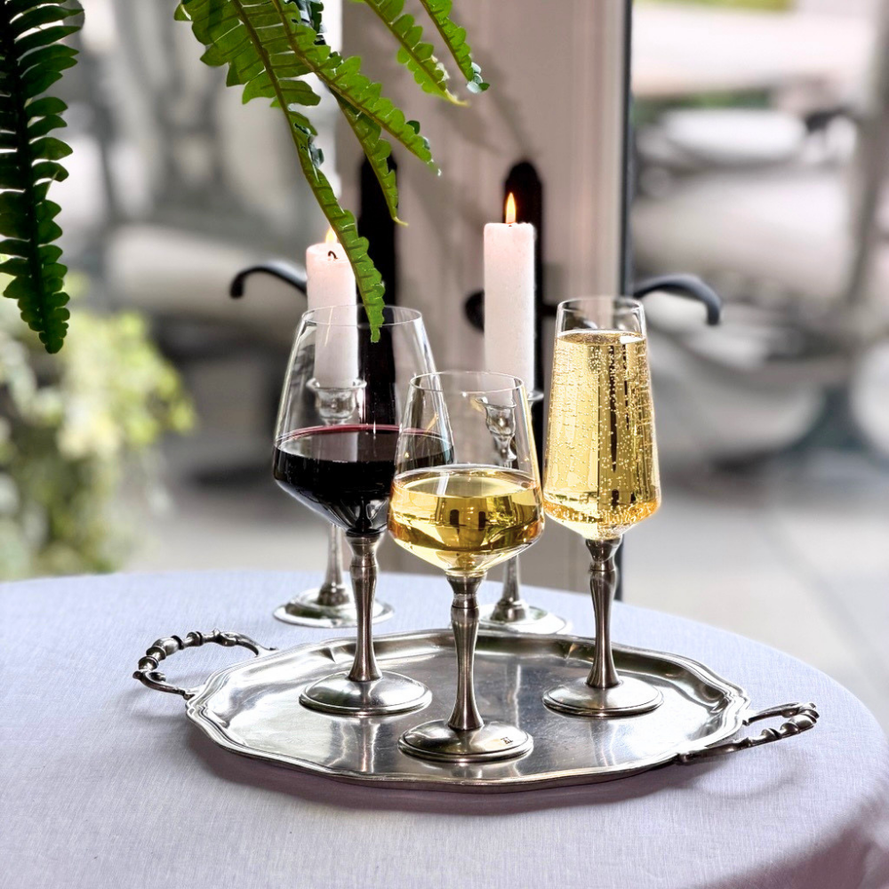 A pewter tray sitting on a table by a window with a Siena Red wine, White wine and Champagne flute with candles behind them