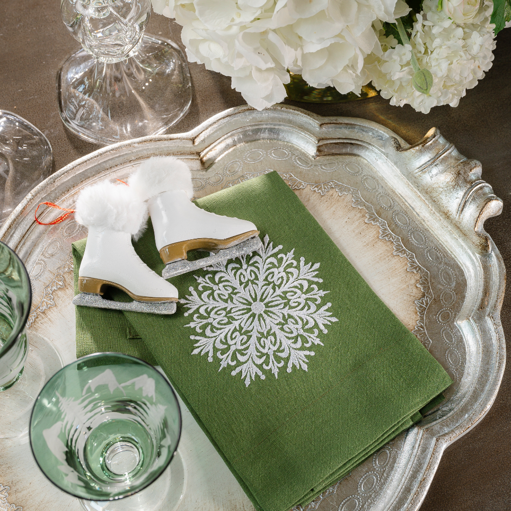 An evergreen linen towel embroidered with a snowflake on a silver Florentino wooden tray with Ice Skate ornaments