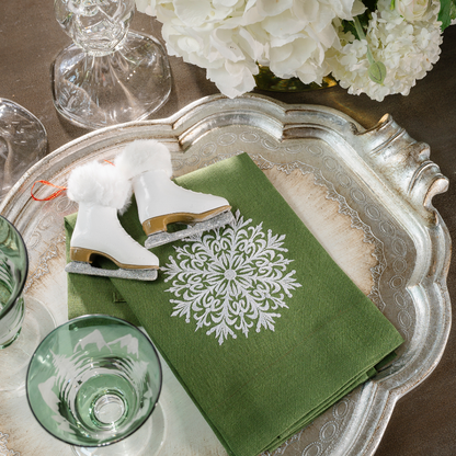 An evergreen linen towel embroidered with a snowflake on a silver Florentino wooden tray with Ice Skate ornaments