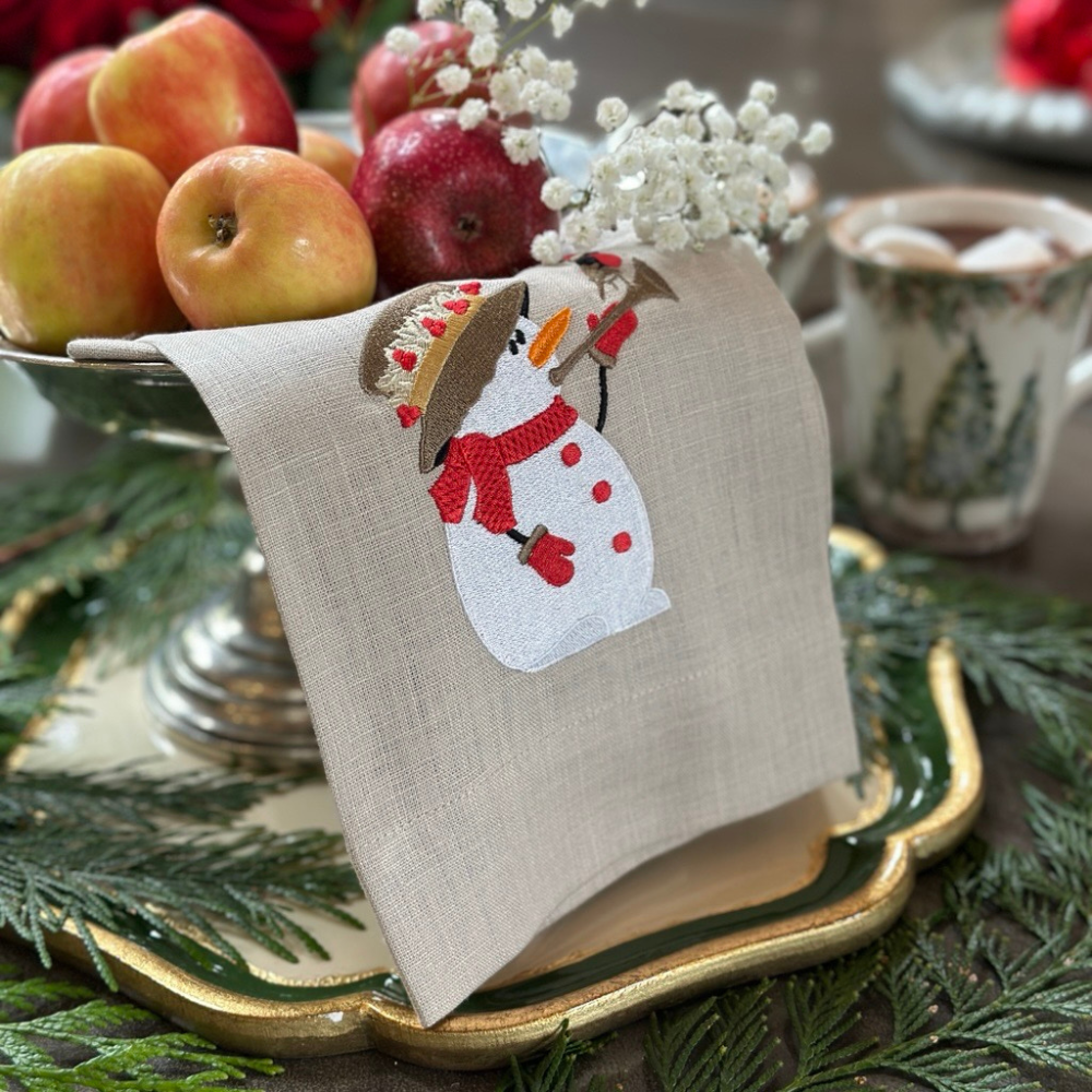 Kitchen towel with snowman playing a horn with a bird design on a festive table setting with apples and coffee.