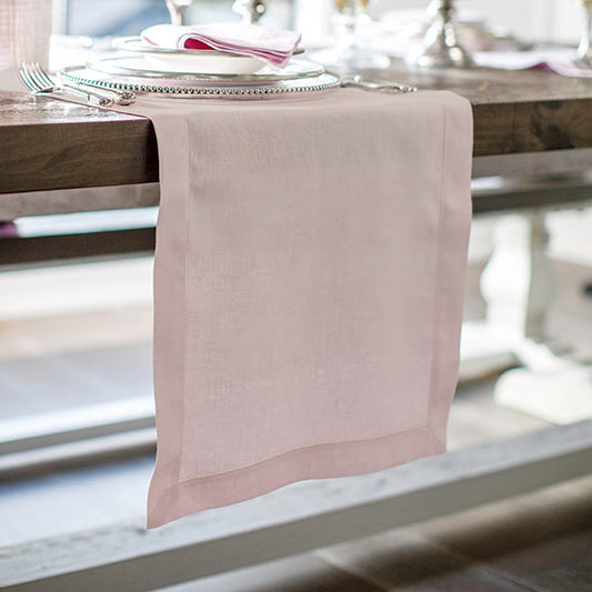 A pink linen table runner with hemstitch detailing, displayed on a table with a floral patterned napkin visible.