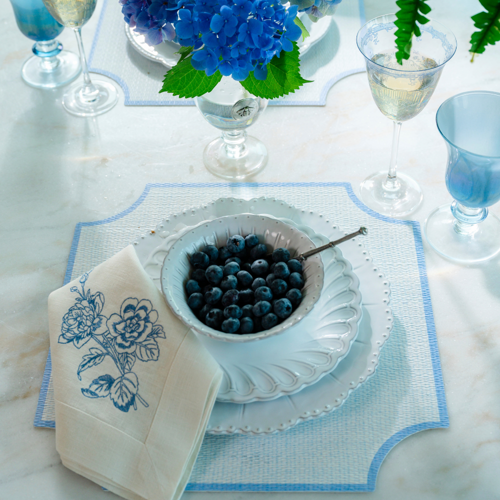 Splendido square placemat with blue trim on a white table with Bella Bianca Plates and Rialto glassware