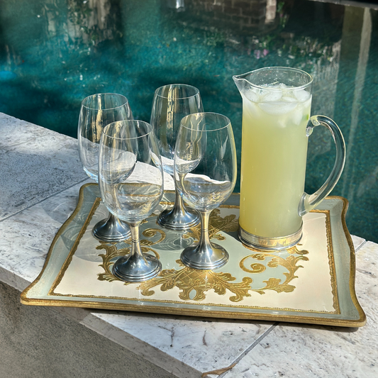 Set of wine glasses and a pitcher of lemonade on an ornate tray by a pool.