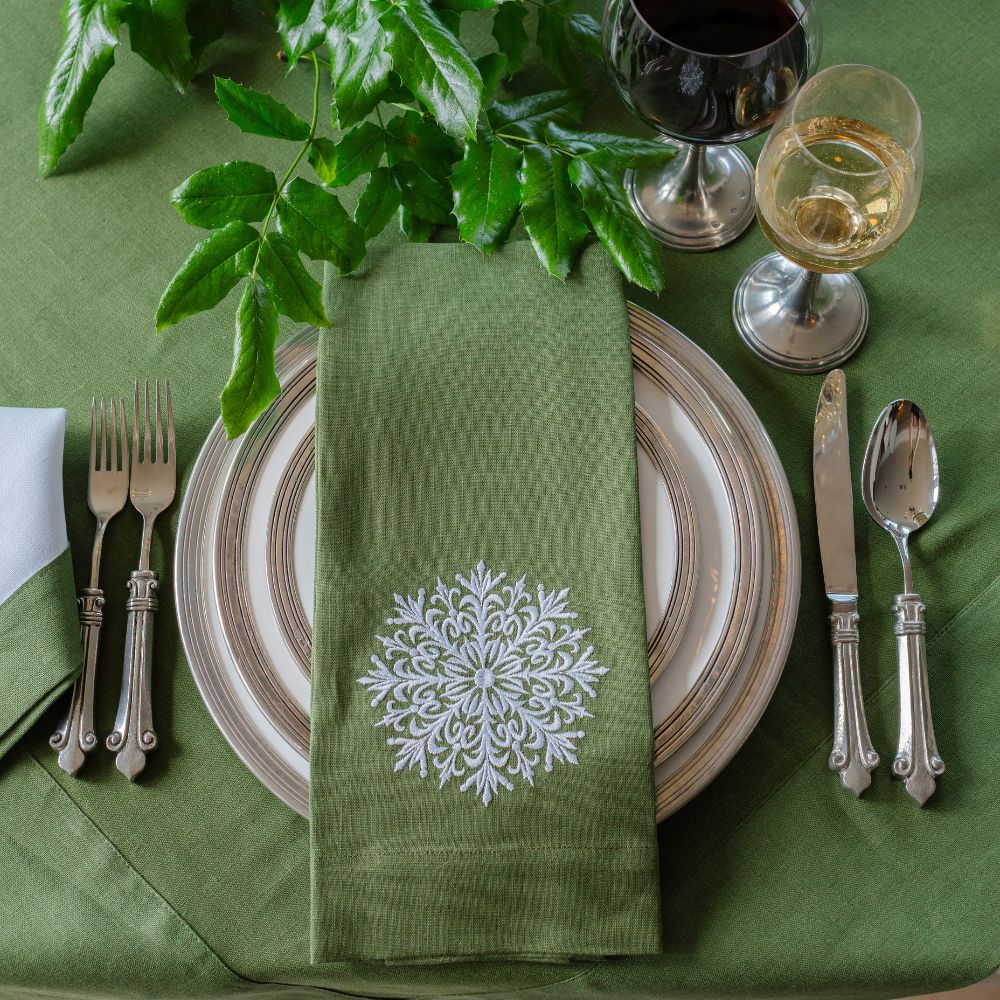 Green table setting with a decorative napkin featuring a snowflake design, plates, cutlery, and glasses on a green tablecloth.