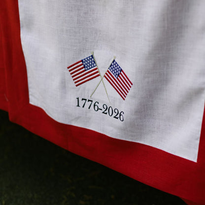 White fabric with embroidered American flags and '1776-2026' text on a red border.