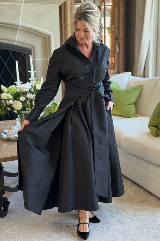 Black taffeta sash overskirt worn over the Peggy button-down dress, showing the full dramatic sweep of the skirt in an elegant living room with a stone fireplace and white floral arrangement