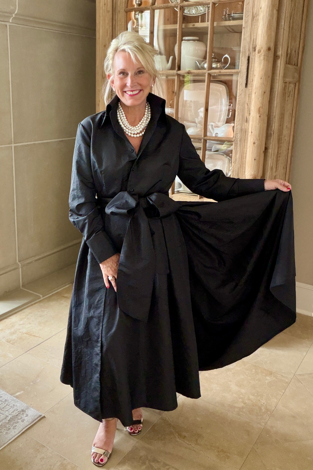 Taffeta sash overskirt in black worn over a black Peggy button-down dress with a bow tie waist, styled with a layered pearl necklace and heeled sandals in an elegant interior with a wooden hutch