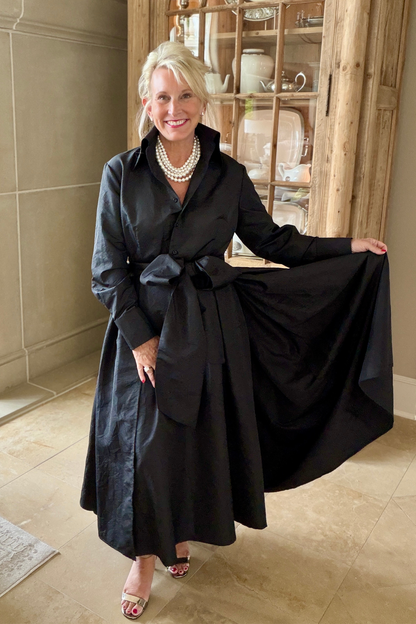 Taffeta sash overskirt in black worn over a black Peggy button-down dress with a bow tie waist, styled with a layered pearl necklace and heeled sandals in an elegant interior with a wooden hutch