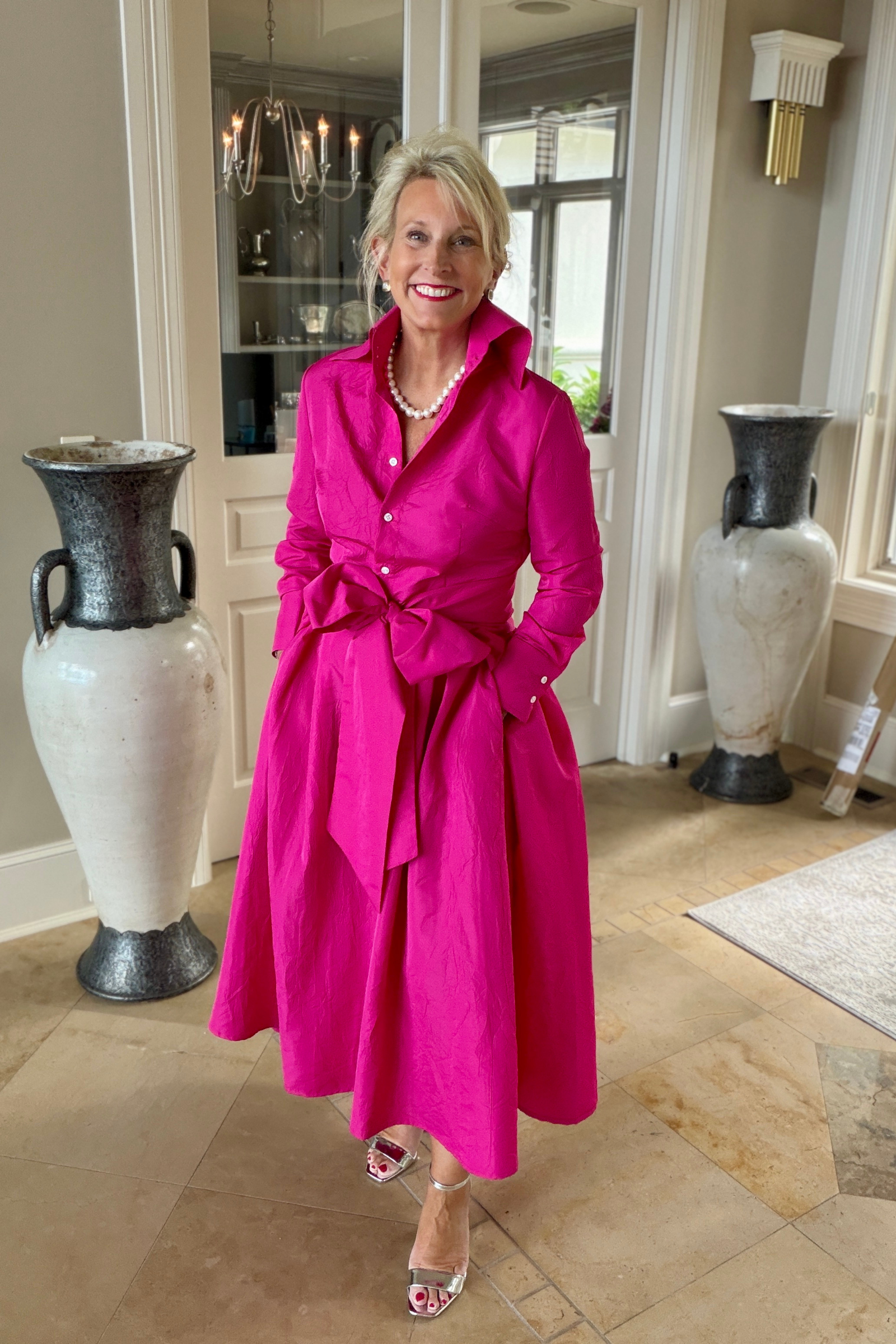 Taffeta sash overskirt in fuchsia worn over a matching fuchsia Peggy button-down dress with a bow tie waist, styled with a pearl necklace and silver heels in an elegant foyer