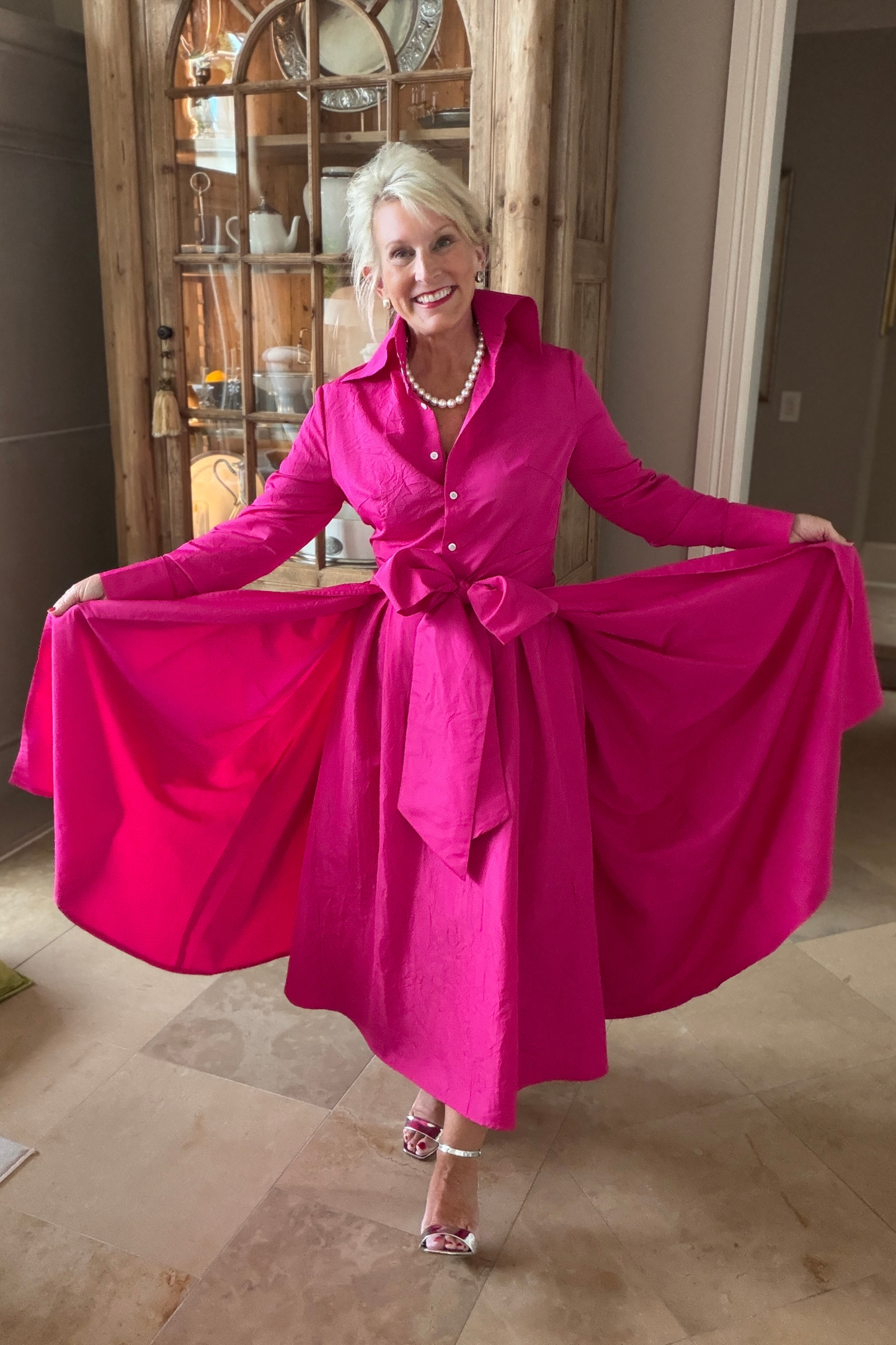 Fuchsia taffeta sash overskirt shown from the sides, held open to display its full voluminous sweep, worn over the Peggy button-down dress with a pearl necklace in a warm interior setting