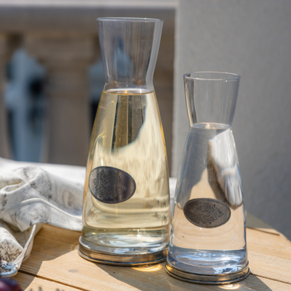 A close up of two glass carafes with pewter base and decorative tab on a wooden table 