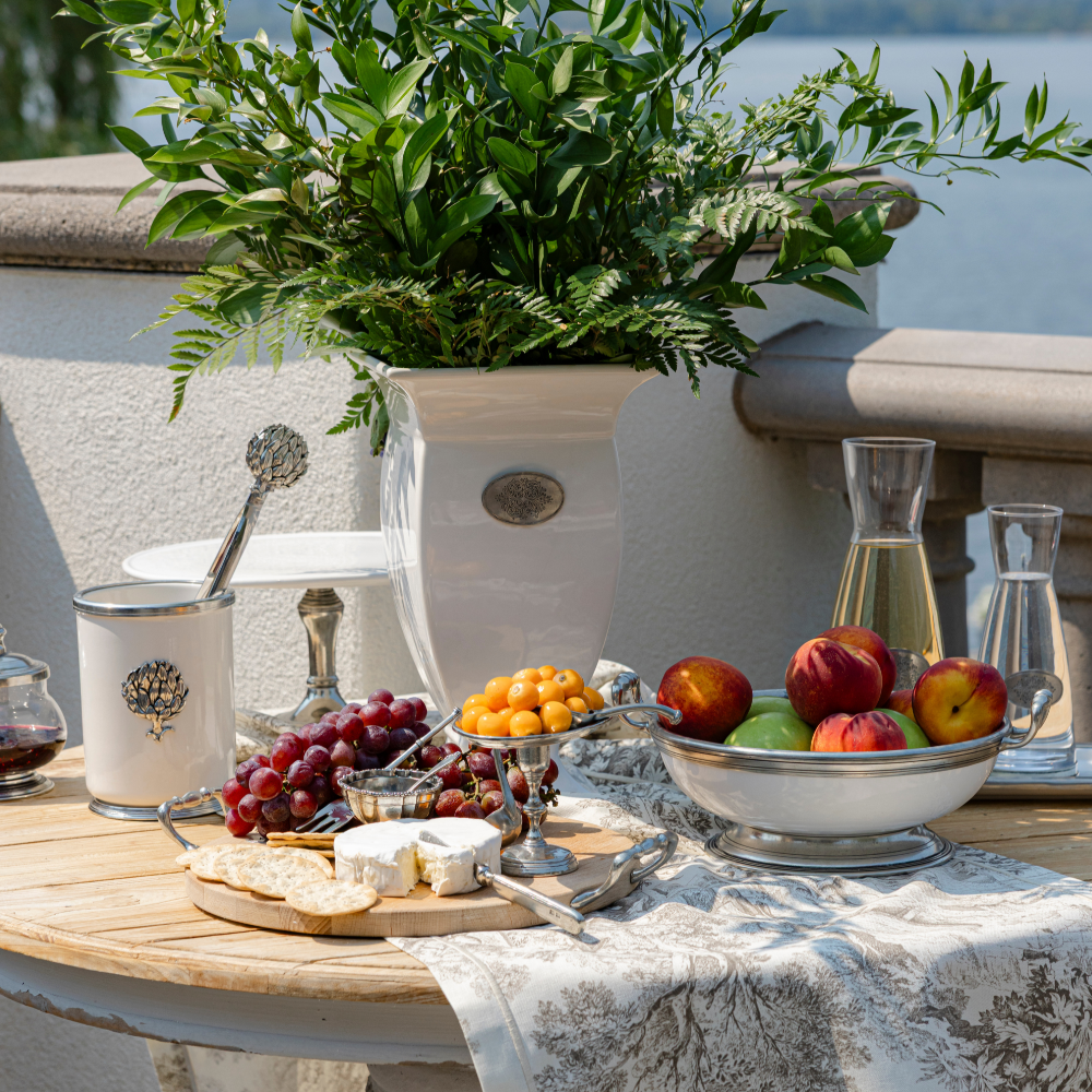 Table set with a Tavola cheese board with grapes and crackers along side a Tuscan medium bowl with peaches and wine and greenery in the background
