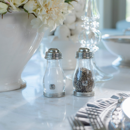 Two clear glass salt and pepper shakers with silver metal caps sit on a white marble tabletop in a styled dining setting, with a white flower vase and glassware in the background.