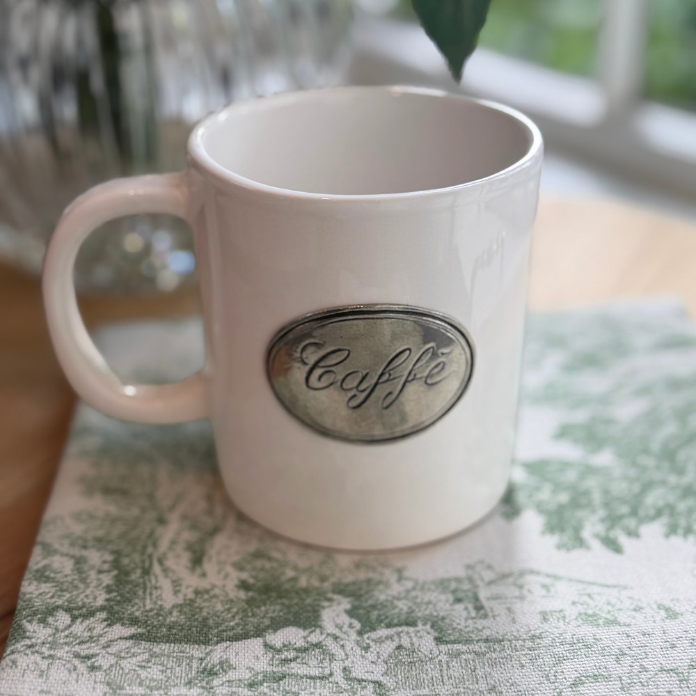 Close up of a Tuscan ceramic with pewter tab that says 'Caffe' sitting on a Montefalco green toile napkin