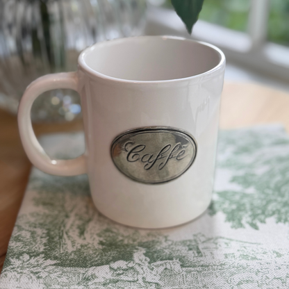 Close up of a Tuscan ceramic with pewter tab that says 'Caffe' sitting on a Montefalco green toile napkin