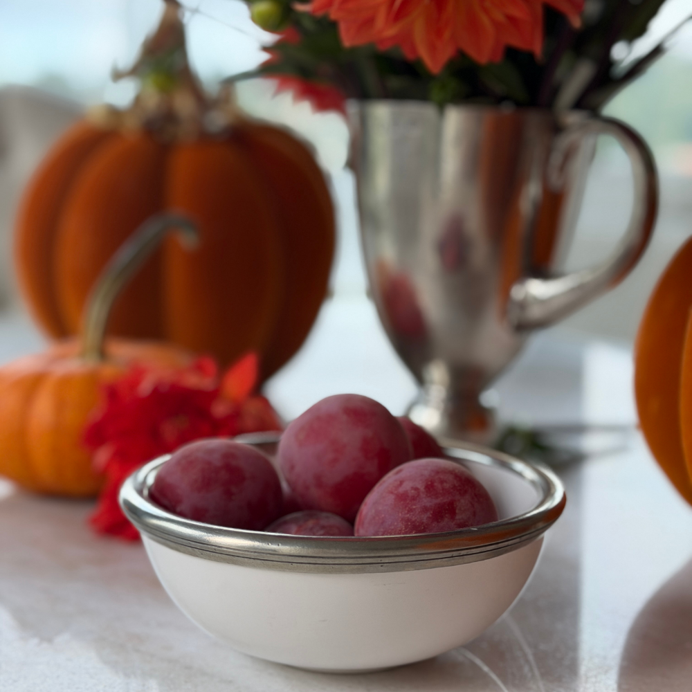A Tuscan ceramic cereal bowl with pewter rim filled with plums on a table with fall decorations