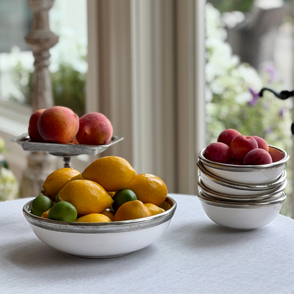 Stack of Arte Italica Tuscan Cereal Bowls with pewter rims filled with fresh nectarines, alongside a Tuscan  medium serving bowl filled with lemons and limes, styled on a white linen tablecloth near a sunny window
