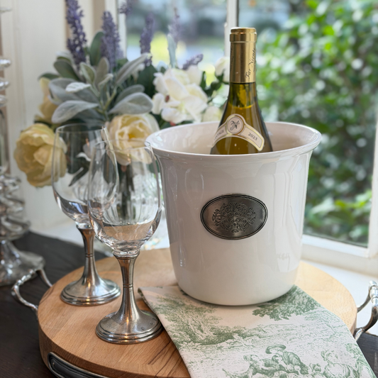 Tuscan ceramic wine bucket with white wine in it sitting on a board with two Verona pewter stem wineglasses with flowers and greenery in the background