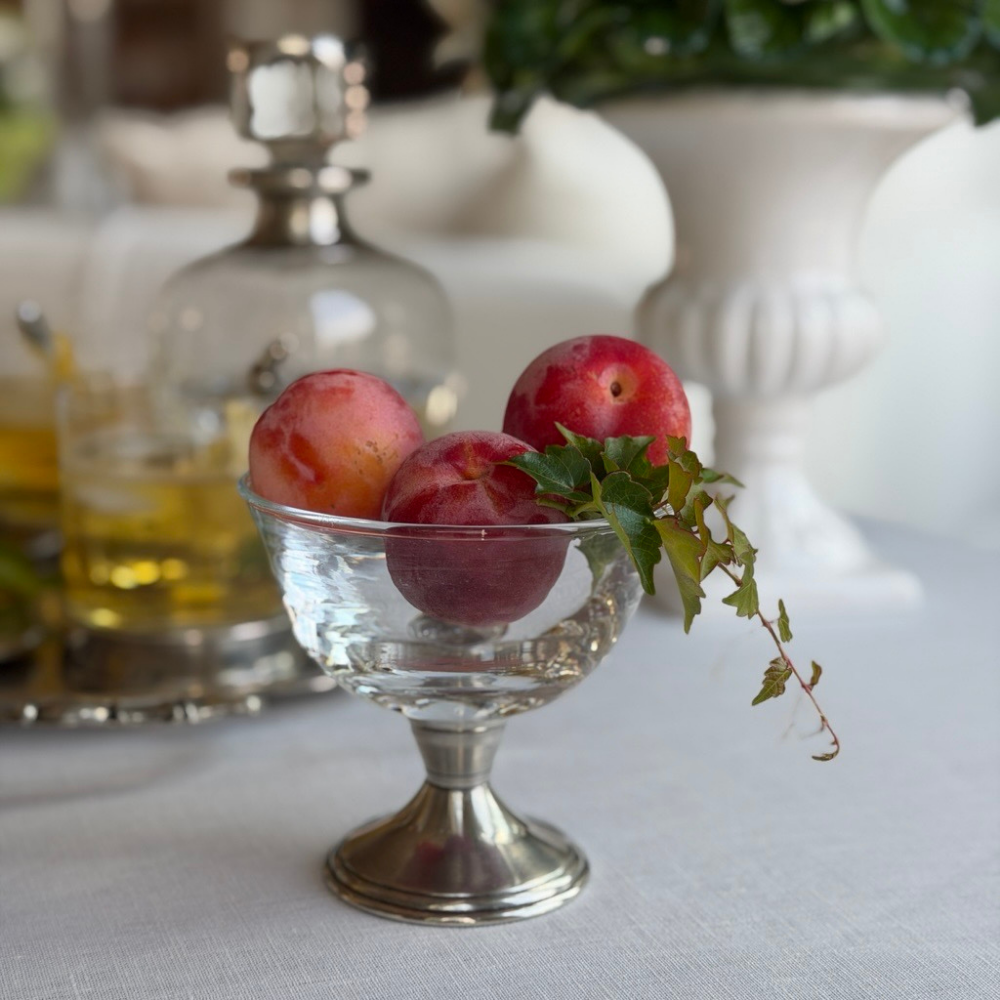 A Tuscan glass compote dish with a pewter base filled with plums on a white table with drinks in the background