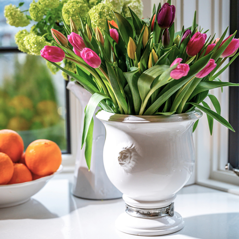 A Tuscan lion ceramic urn with a pewter accent with Pink tulips on a counter with oranges in a bowl