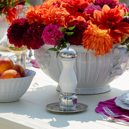 A white table with bella bianca dinnerware and garden urn with orange and magenta dahalias with a Tuscan pepper ginder