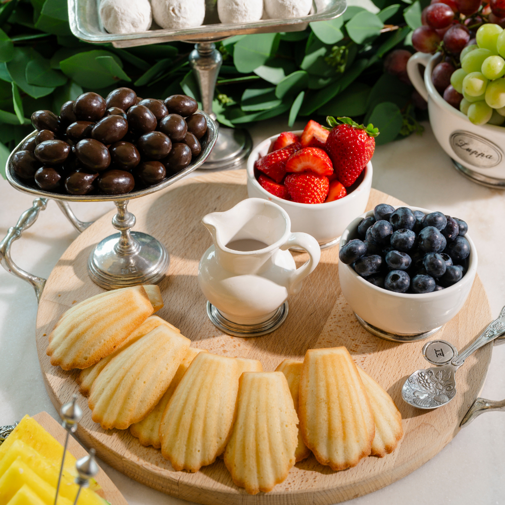 A dessert charcuterie board with strawberries, blueberries, chocolate almonds and madeleines on a wood board with a pitcher of sauce