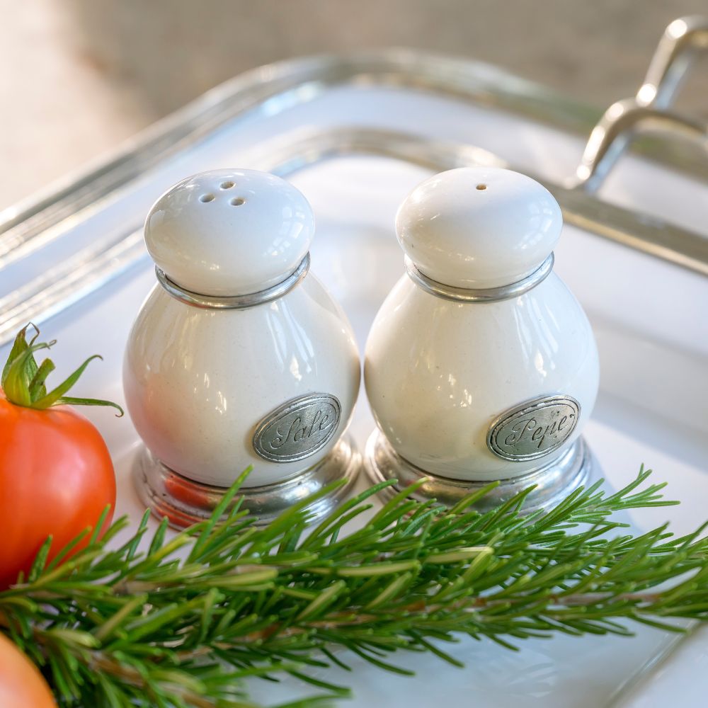 Two white ceramic salt and pepper shakers on a Tuscan tray with a tomato and rosemary sprig.