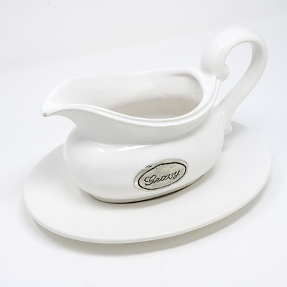 White ceramic gravy boat with pewter tab on a white plate against a plain background