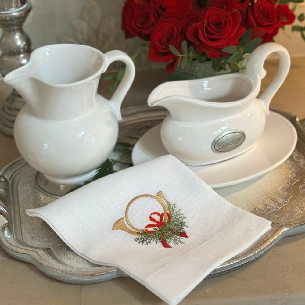 White ceramic Tuscan pitcher and gravy boat with saucer on a Florentino silver tray with a decorative napkin featuring a Christmas Horn with bow.
