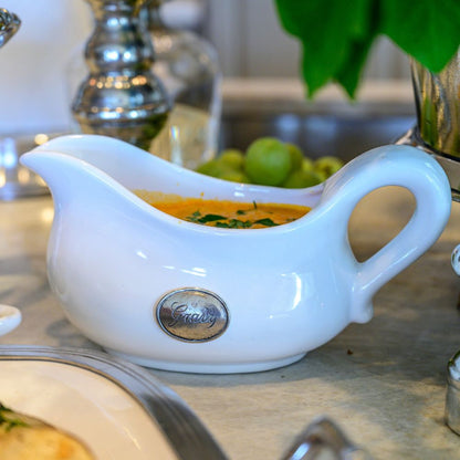 White gravy boat with a pewter gravy label on a table with food in the background