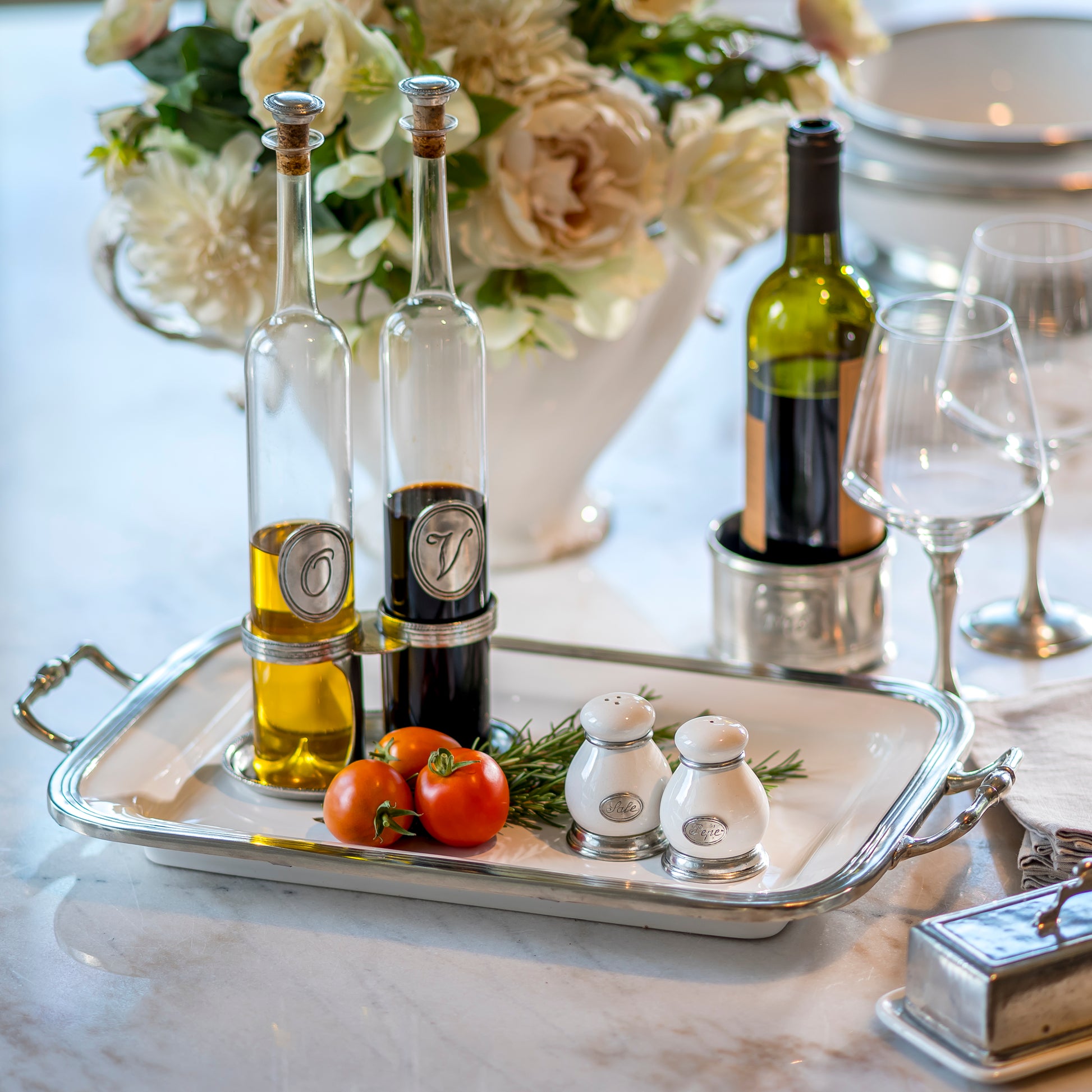 Bottles of olive oil and vinegar on a tray with tomatoes and Tuscan salt and pepper shakers, set against a floral background.