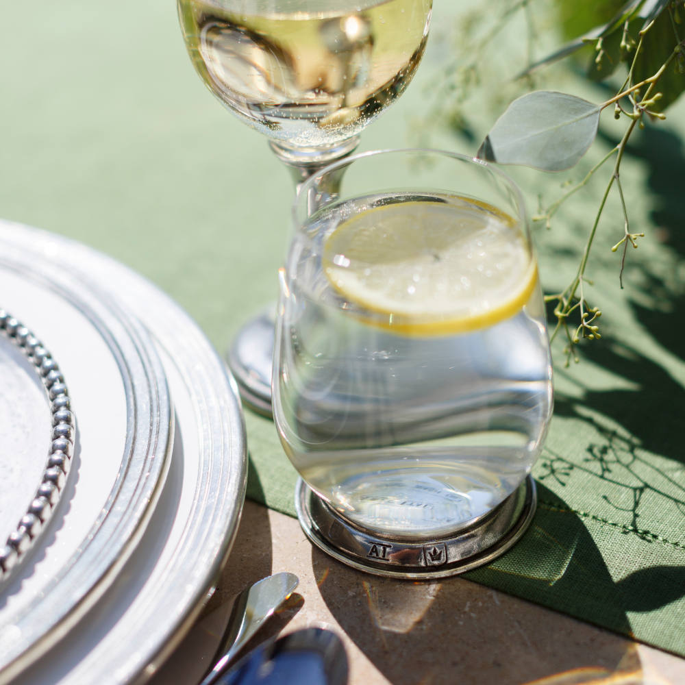 A Verona glass tumbler with a pewter base filled with water and a lemon on a green linen runner next to Tuscan plates