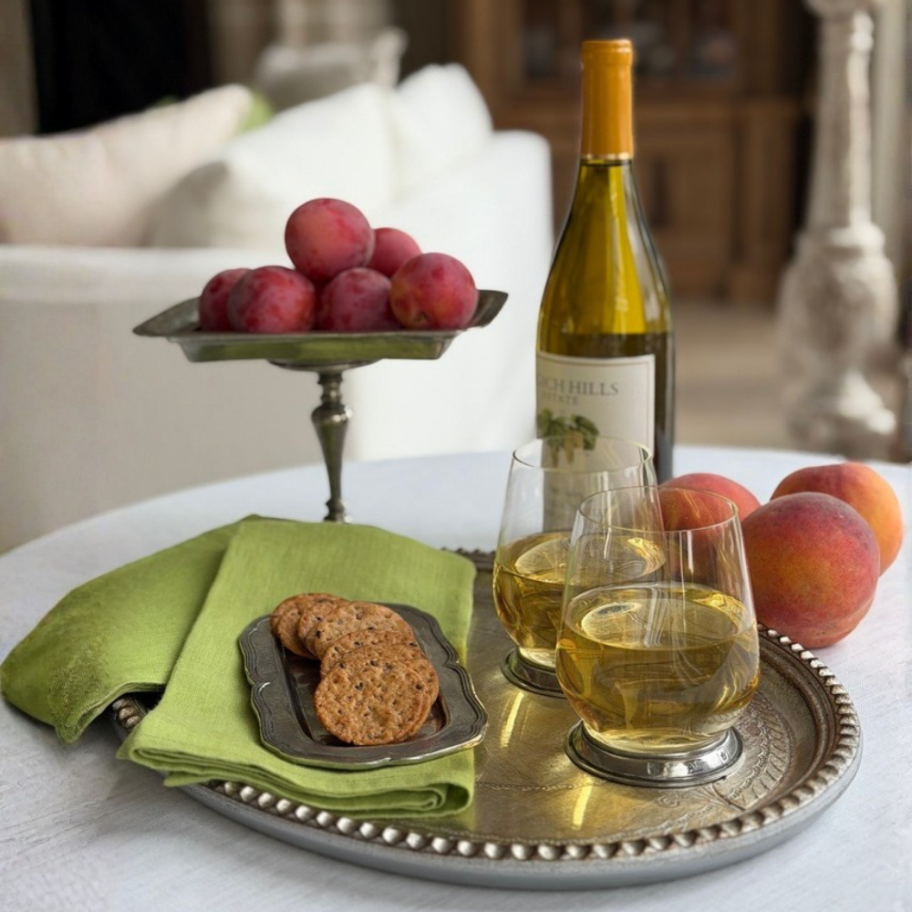 Two Verona glass tumblers with pewter base on a silver tray filled with white wine, with peaches and plums, citron linen napkins on a table