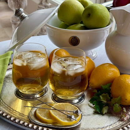 Two Verona tumblers filled with ices Lemonade on a tray with sliced and whole lemons and a bowl with green apples behind it